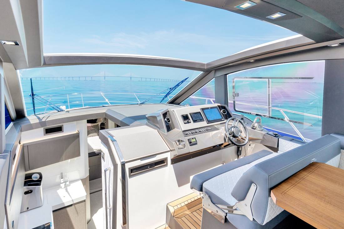 Luxury motor yacht helm with modern navigation consoles, stainless-steel wheel, quilted leather bench and teak decking, panoramic windshield framing clear blue sea and distant bridge under a sunny sky.