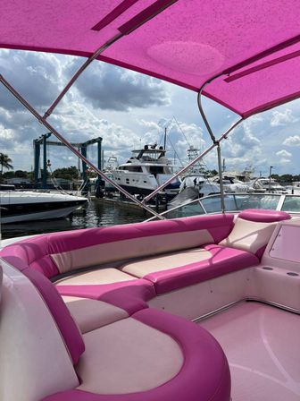 Vibrant pink-and-white lounge seating and matching pink bimini on a leisure boat docked at a marina, with large yachts and a partly-cloudy sky in the background.