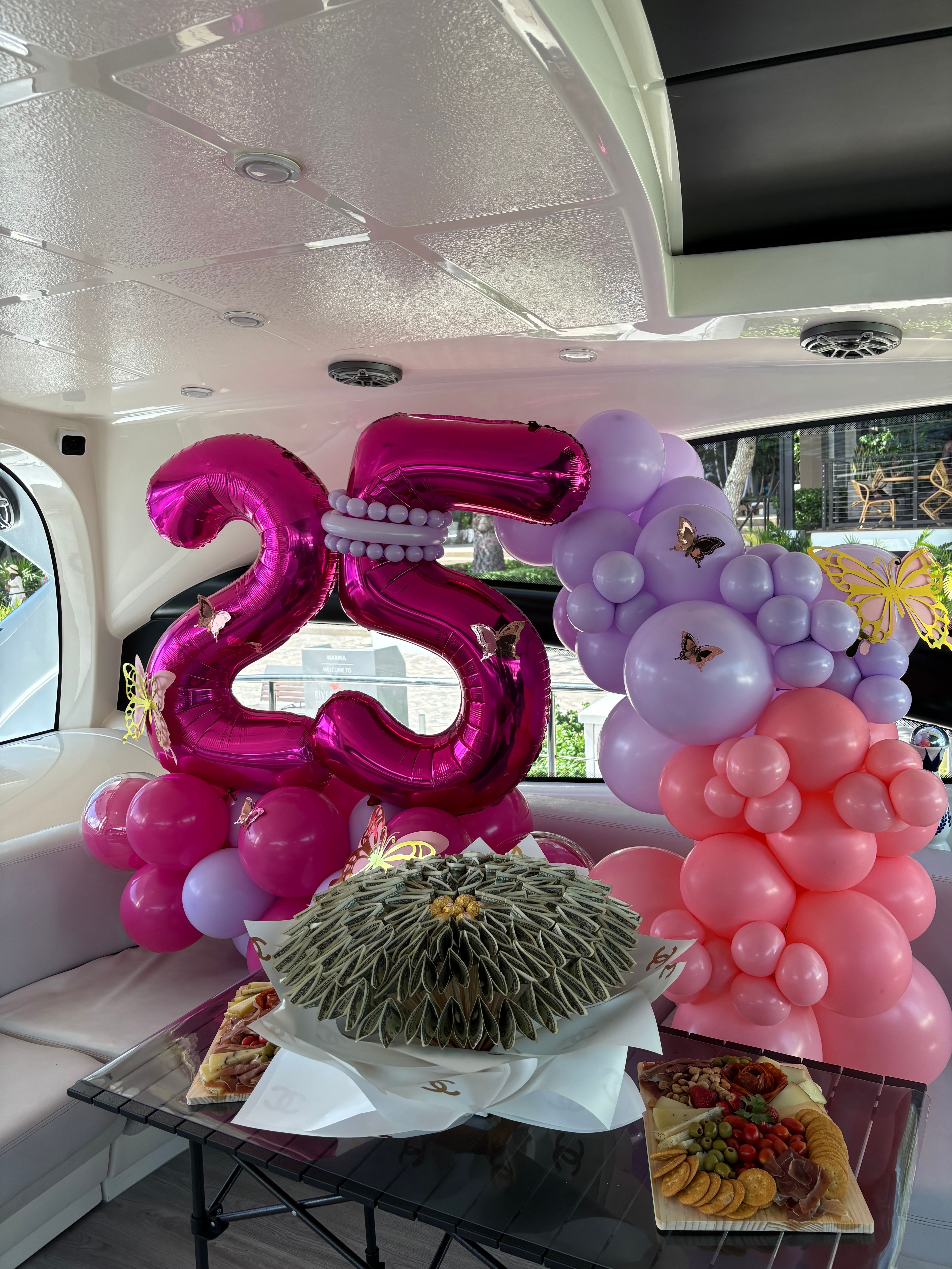 Luxury yacht interior decorated for a 25th birthday with large metallic pink "25" balloons, pink and lavender balloon garland with butterfly accents, a folded-bill money bouquet centerpiece and a charcuterie board with cheeses, crackers and fruit.