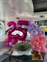 Luxury yacht interior decorated for a 25th birthday with large metallic pink "25" balloons, pink and lavender balloon garland with butterfly accents, a folded-bill money bouquet centerpiece and a charcuterie board with cheeses, crackers and fruit.