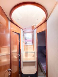 Sunlit luxury yacht interior corridor with warm wooden trim and steps leading up to a pink salon and distant city skyline view.