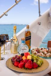Yacht brunch table with spirits and champagne, elegant gold-rimmed glasses, colorful fruit platter and savory skewers, set against a turquoise sea.