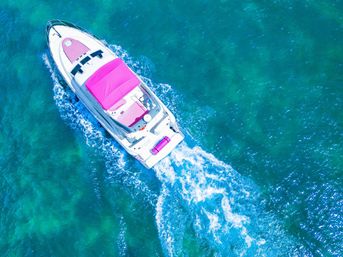 Aerial view of a white luxury yacht with a pink sunshade cruising clear turquoise ocean, leaving a foamy wake.
