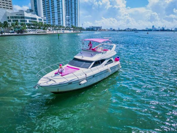 White motor yacht with pink cushions and canopy, two people in pink aboard, cruising a turquoise bay past a palm-lined urban waterfront and high‑rise buildings on a sunny day.