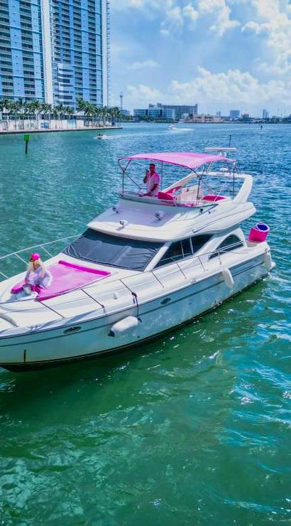 White motor yacht with pink cushions and canopy, two people in pink aboard, cruising a turquoise bay past a palm-lined urban waterfront and high‑rise buildings on a sunny day.