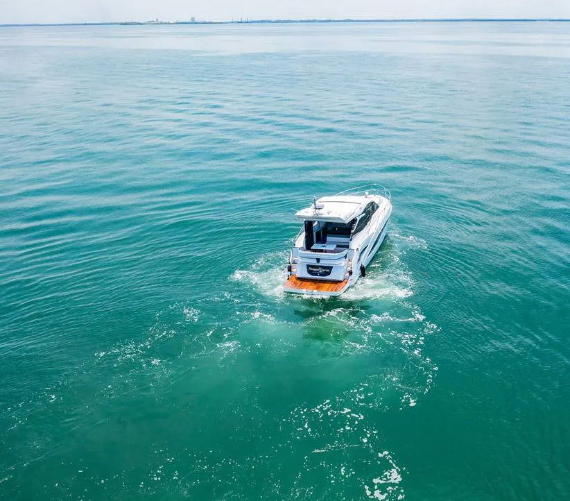 Aerial view of a white motorboat cruising on calm turquoise open water, leaving a foamy wake behind toward a distant flat horizon.