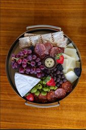 Round gourmet charcuterie platter on a wooden table with a brie wedge, sliced salami, cubed cheese, crisp crackers, red grapes, green olives, strawberries, blackberries, chocolate-covered bites, and a small jar of jam.