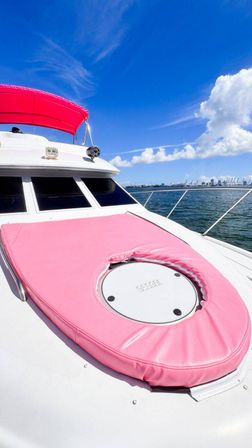 Yacht bow with bright pink sun pad and matching canopy on a white deck, vivid blue sky and coastal harbor skyline on the horizon