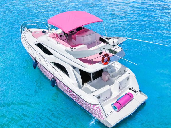 Aerial view of a white motor yacht with bright pink canopy and accents, anchored in crystal-clear turquoise tropical water.