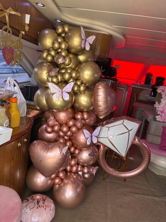 Rose-gold and gold balloon garland with butterfly cutouts, heart and diamond-ring foil balloons arranged inside a yacht cabin for a bachelorette-style celebration