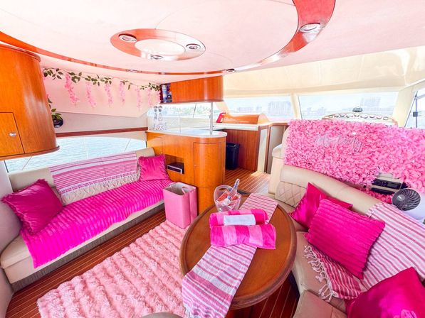 Luxury yacht interior salon with playful hot-pink decor — plush pink rugs, throws and cushions on cream leather seating, wooden coffee table with striped towels and ice bucket, floral garland and waterfront view through large windows.