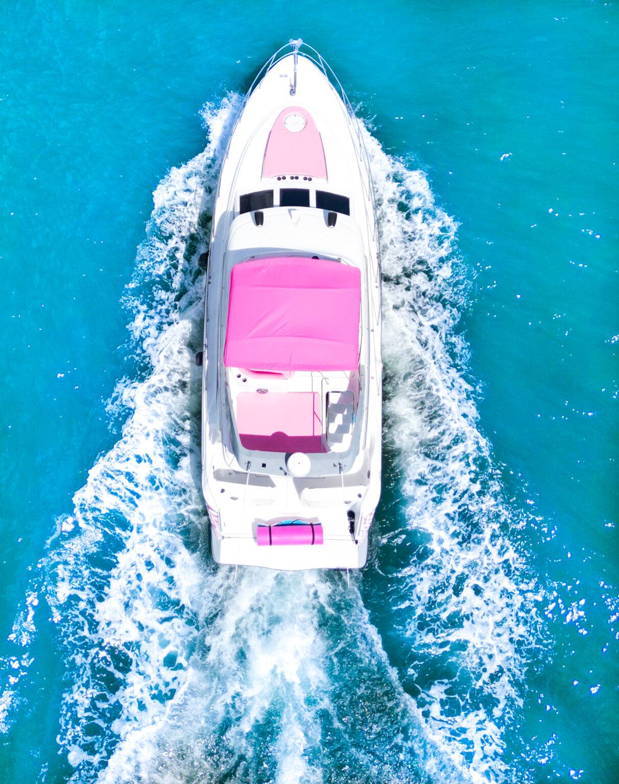 Aerial drone view of a white luxury yacht with a bright pink canopy cruising through clear turquoise water, trailing a foamy wake
