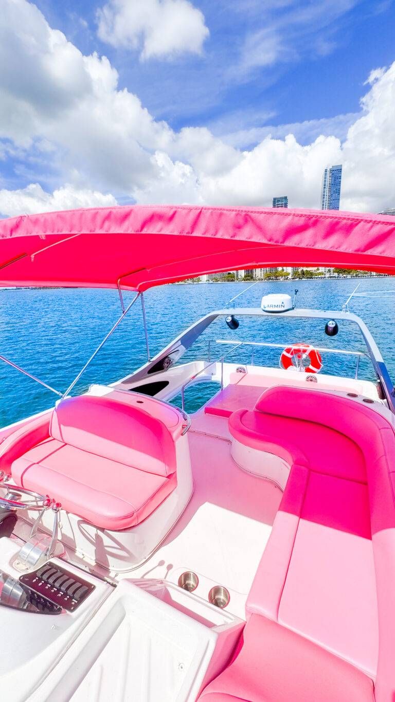 Bright pink speedboat interior with curved cushioned seats and canopy on a turquoise bay, waterfront city skyline and fluffy clouds under a blue sky