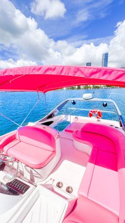 Bright pink speedboat interior with curved cushioned seats and canopy on a turquoise bay, waterfront city skyline and fluffy clouds under a blue sky