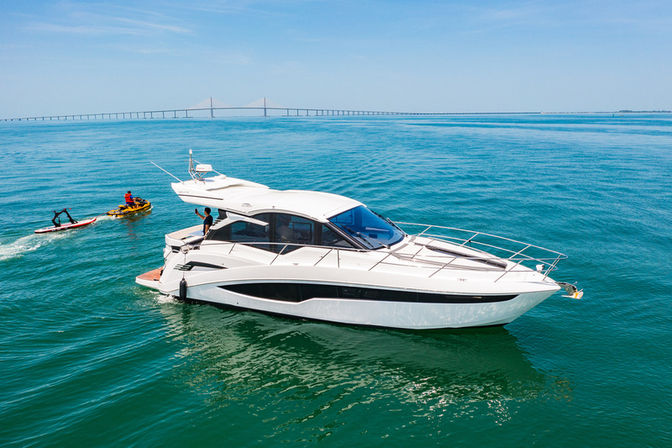 White luxury motor yacht on turquoise coastal waters near a long bridge, with people riding a jet ski and paddleboard under a clear blue sky.