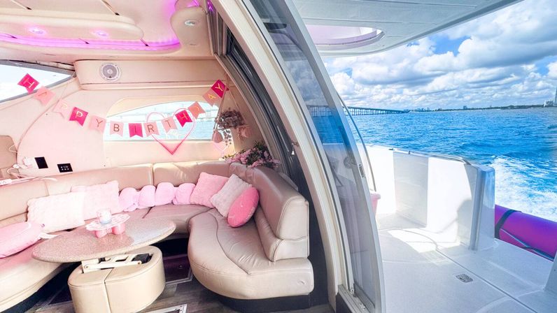 Pink-themed luxury yacht interior with plush curved seating and birthday banner, party-ready table opening onto sunny blue ocean and a distant bridge over coastal waters.
