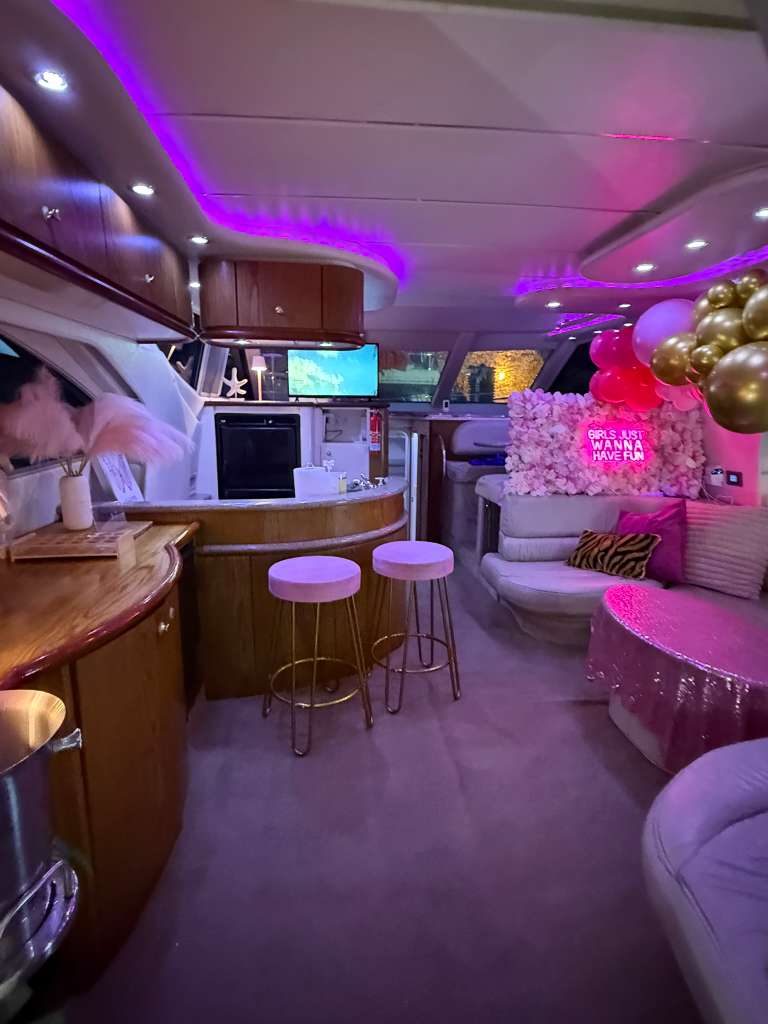 Luxury yacht party interior with pink and purple LED lighting, wooden bar and two pink stools, floral neon backdrop saying 'Girls Just Wanna Have Fun', gold balloons and plush seating.