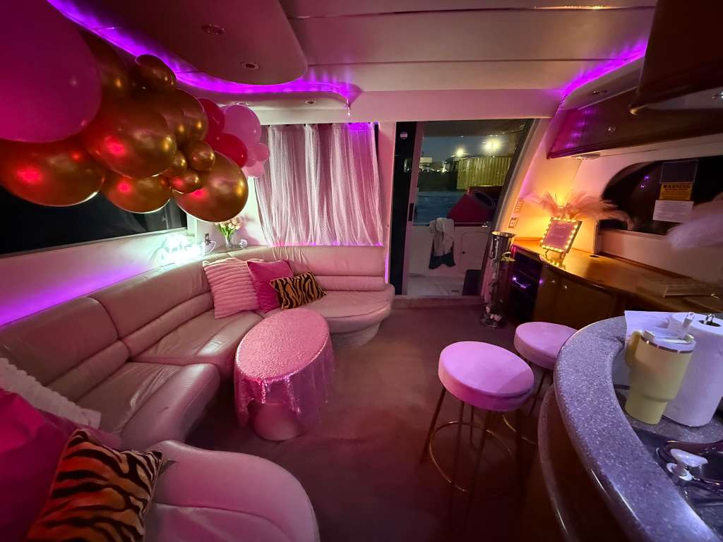 Pink-and-purple lit luxury yacht lounge with curved leather sofa, gold and pink balloons, sequin-covered cocktail table, bar stools and a marina view through the sliding door