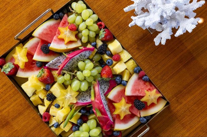 Colorful tropical fruit platter with watermelon slices, dragon fruit, pineapple cubes, green grapes, starfruit, strawberries, blueberries and blackberries on a wooden table beside a white coral centerpiece