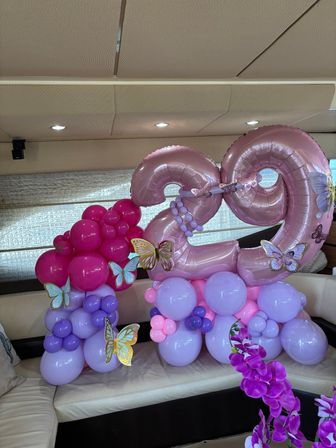 Festive balloon display with metallic pink "29" formed by clusters of hot pink, lavender and purple balloons, butterfly accents and purple orchids on a cream sofa inside a cabin — birthday decoration.