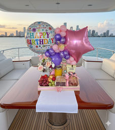 Birthday brunch display on a yacht table: colorful "Happy Birthday" foil balloon with pink star and purple balloon garland, flowers, chocolate-dipped strawberries, fresh fruit, pastries and juice, set against a waterfront skyline at sunset.
