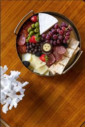 Top-down view of a round charcuterie board on a warm wooden table featuring a wedge of brie, cubed white cheese, sliced salami, red grapes, strawberries and blackberries, green olives, crackers and a small jar of spread — a colorful gourmet cheese platter.