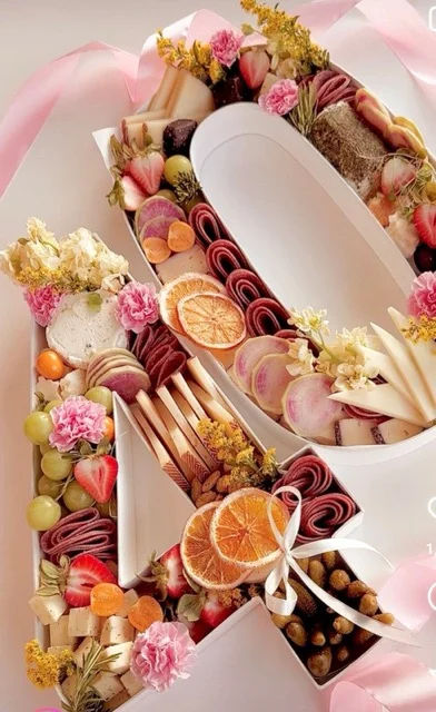 Pink-themed letter-shaped charcuterie gift box with rolled cured meats, assorted cheeses, dried orange slices, strawberries, grapes, nuts, pickles, edible flowers and a pink ribbon
