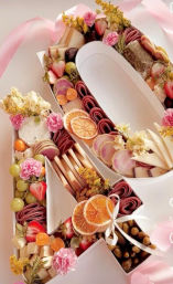 Pink-themed letter-shaped charcuterie gift box with rolled cured meats, assorted cheeses, dried orange slices, strawberries, grapes, nuts, pickles, edible flowers and a pink ribbon