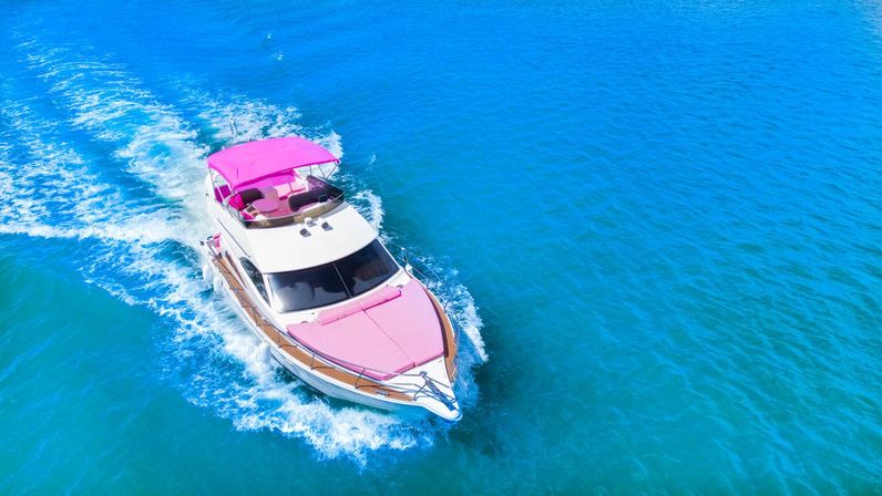 Aerial view of a white motor yacht with bright pink canopy and cushions cruising through clear turquoise ocean water, leaving a foamy white wake.