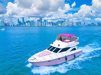 White-and-pink luxury motor yacht with pink canopy speeding across turquoise water in front of the Miami skyline under blue sky and puffy clouds.