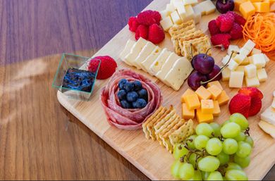 Colorful charcuterie board on a wooden table with green grapes, cubed cheddar and white cheeses, a salami rose filled with blueberries, assorted crackers, cherries, strawberries, and a small dish of jam.