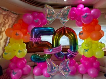 Festive indoor 50th birthday balloon display with rainbow foil '50' numbers framed by hot pink, orange and yellow balloon columns, iridescent bow balloons and butterfly accents