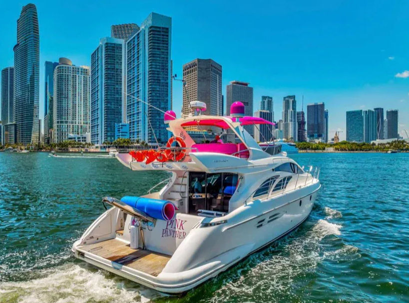 Playful white luxury yacht with vibrant pink canopy cruising the Miami waterfront past a modern downtown skyline under a bright blue sky
