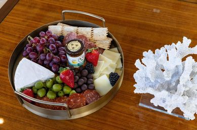 Round metal charcuterie tray on a polished wooden table with brie wedge, sliced cheese, salami, red grapes, green olives, strawberries, blackberries, crackers and a small jar of honey, beside white coral décor.