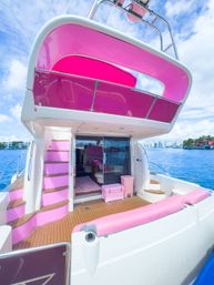 Pink-accented luxury yacht stern with teak deck, pink cushions and canopy, curved steps to upper deck, moored near a coastal skyline under a bright blue sky