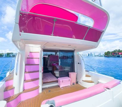 Pink-accented luxury yacht stern with teak deck, pink cushions and canopy, curved steps to upper deck, moored near a coastal skyline under a bright blue sky
