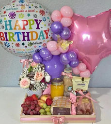 Birthday gift tray with a colorful "Happy Birthday" balloon, pink star and purple balloon cluster, floral chocolate bouquet, fresh fruit, mason-jar orange juice, pastries and wrapped treats tied with pink ribbons.