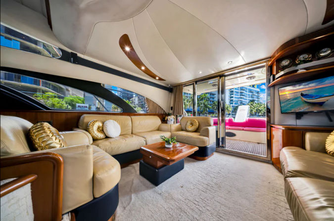 Luxury yacht salon with beige leather sofas, wooden accents and coffee table, panoramic windows and sliding glass doors opening to a sun deck with bright seating and a marina view of palm trees and modern waterfront high-rises