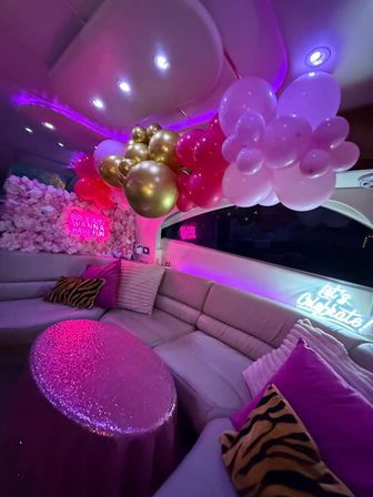 Pink-lit party lounge with gold and pink balloon garland, floral backdrop, neon signs, plush curved seating, zebra-print pillows and sequin-covered table — bachelorette or girls' night celebration setup