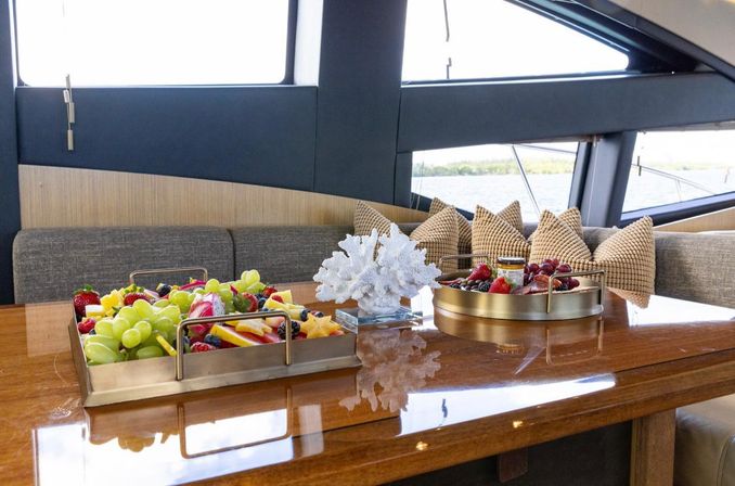 Luxury yacht dining area with polished wood table, gold trays of colorful fresh fruit, decorative white coral centerpiece, textured beige cushions and water view through large windows.