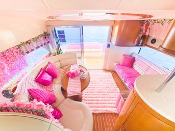 Bright pink-themed luxury yacht interior — curved cream leather salon with fuchsia pillows, striped pink throws, fluffy pink rug, wooden table and floral garlands, sliding glass doors opening to ocean view