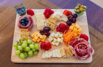 Colorful charcuterie board on a wooden serving board featuring assorted cheeses (sliced and cubed), crackers, green grapes, strawberries, raspberries, cherries, blueberries tucked into salami roses, shredded carrots, a small cup of olives and a cup of jam.