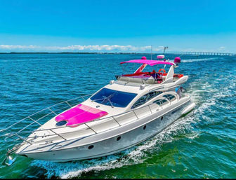 Luxury white motor yacht with bright pink canopy and deck accents cruising on turquoise ocean water under a clear blue sky near a long coastal bridge.
