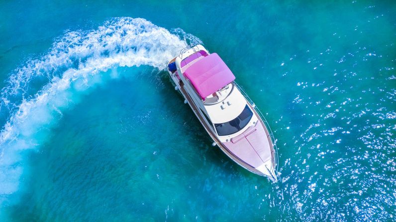 Aerial drone view of a white and pink luxury yacht carving a curved wake through sparkling turquoise ocean water