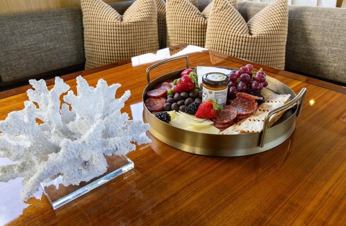 Round brass charcuterie tray with salami, sliced cheese, strawberries, blackberries, red grapes, chocolate-covered nuts, crackers and jam jar on a glossy wooden table next to white coral decor and beige textured pillows in a cozy seating nook.