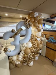 Gold, white and tan balloon garland forming large '30' number balloons with metallic floral accents and small gold butterflies, displayed inside a yacht salon as a party decoration.