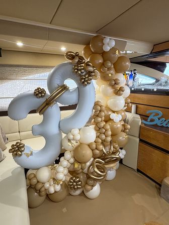 Gold, white and tan balloon garland forming large '30' number balloons with metallic floral accents and small gold butterflies, displayed inside a yacht salon as a party decoration.