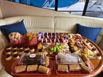 Colorful charcuterie and dessert spread on a round wooden table inside a yacht salon — cheeses, crackers, nuts, grapes and strawberries, assorted pastries, cookies, mini puddings and packaged cakes arranged on platters.