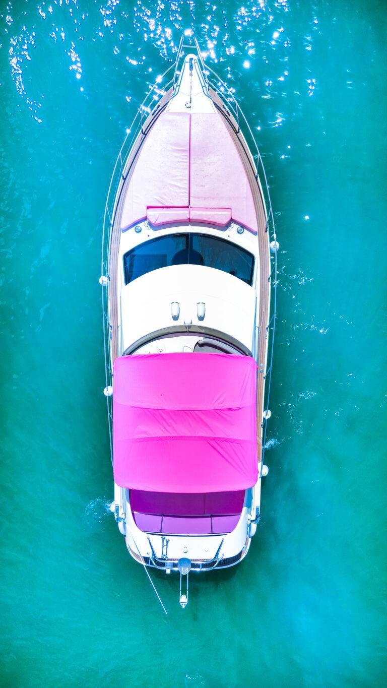 Aerial drone view of a white yacht with a vibrant pink canopy floating on turquoise water with sparkling sunlight reflections
