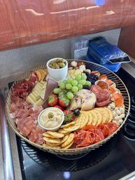 Colorful round charcuterie board on a kitchen countertop with mixed cheeses, green grapes, strawberries, olives, crackers, hummus, nuts, jam and assorted cured meats — gourmet appetizer platter.